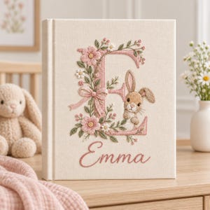 May include: A cream-colored fabric-covered book featuring an embroidered design of the letter E in pink, decorated with floral vines, a small bow, and a brown bunny. The name Emma is embroidered in pink cursive script below the letter.
