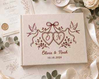 May include: A rectangular beige wedding guest book featuring dark red embroidery of two doves holding ribbons connected to two hearts with the letters O and N inside. Below the hearts, the names Olivia & Noah and the date 10.18.2026 are embroidered.