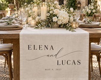 Könnte beinhalten: Ein beigefarbener Tischläufer aus Leinen mit dem aufgedruckten schwarzen Text ELENA and LUCAS und dem Datum MAY 9, 2027. Der Läufer liegt auf einem Holztisch, der mit weißen Blumenarrangements, Kerzen und Gläsern gedeckt ist.