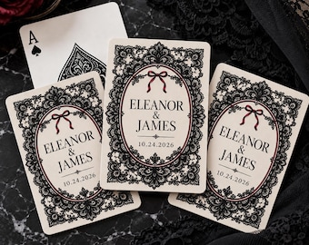 Könnte beinhalten: Drei personalisierte Spielkarten mit einer komplexen schwarzen Spitzenbordüre, einem dunkelroten ovalen Rahmen und einer kleinen roten Schleife. Der mittlere Text lautet Eleanor & James mit dem Datum 10.24.2026. Ein Pik-Ass ist teilweise sichtbar.