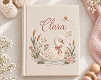 May include: A cream-coloured fabric-covered book featuring an embroidered white swan with a pink bow, cattails, and flowers. The name Clara is embroidered in pink script above the swan, accompanied by a small dragonfly and floral accents.