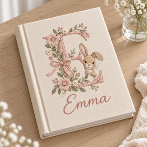 May include: A cream-colored hardcover book featuring intricate embroidery on the front cover. The design includes a large letter E decorated with pink flowers, green leaves, a small bow, and a brown rabbit. The name Emma is embroidered below.