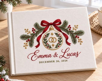 May include: A white fabric-covered photo album featuring an embroidered design of a Christmas ornament with two gold rings, green pine branches, and a red bow. The text reads Emma & Lucas and DECEMBER 26, 2026 in red and gold embroidery.