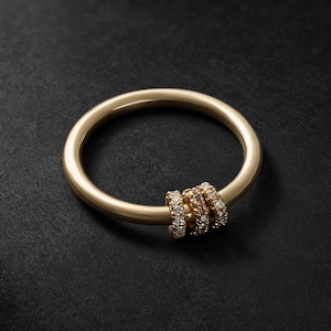 Peut inclure: Un bracelet jonc doré avec trois petits anneaux ornés de petits pierres précieuses transparentes. Le bracelet est posé sur un fond sombre, mettant en valeur le design et la couleur du bijou.