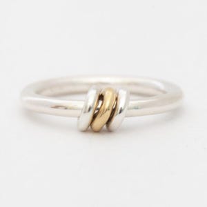 Peut inclure: Une bague en argent avec une bande épaisse, ornée de trois anneaux connectés : deux argentés et un doré. La bague est posée sur un fond blanc, soulignant son design simple mais élégant. Un bijou.