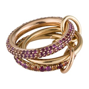 Op de afbeelding: Een set gouden ringen die in elkaar grijpen. Eén ring is bezet met kleine, ronde, paarse edelstenen. De andere ringen zijn effen goud. De ringen zijn ontworpen om samen als set te worden gedragen.