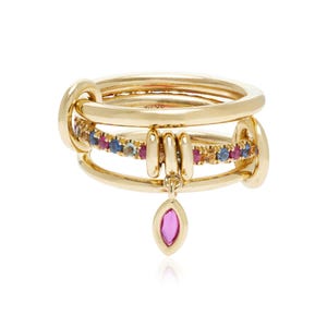 May include: A gold ring featuring multiple stacked bands connected by small loops. One band is set with small multicoloured gemstones, and a single marquise-cut pink gemstone hangs as a charm from the centre of the ring structure.