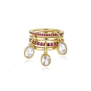 May include: A gold multi-band ring featuring two bands encrusted with small red stones and two plain gold bands. Three oval-shaped clear white gemstones hang from the rings as charms, each set in a gold frame with a beaded edge detail.