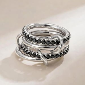 May include: A collection of silver rings featuring black gemstones. The rings are interlocked, forming a distinctive, contemporary design. The gemstones are arranged in a line around the top of the rings.