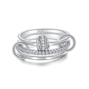 May include: A silver-toned ring featuring a multi-band design with a central row of small clear stones. The ring includes a prominent vertical cylindrical element encrusted with clear stones and is stamped with the text S925 on the inner band.