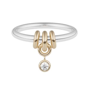 May include: A silver band ring featuring three gold-toned rings suspended from the top, with a single gold-toned charm dangling below. The charm consists of a small circular gold frame holding a single clear, round-cut gemstone.