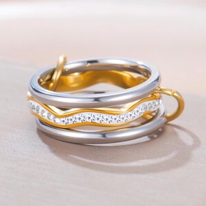 Puede incluir: Un conjunto de anillos apilables. El conjunto incluye una banda plateada, una banda dorada y una banda dorada con cristales transparentes. Los anillos están entrelazados y tienen un acabado brillante. Las joyas se exhiben sobre una superficie neutra.