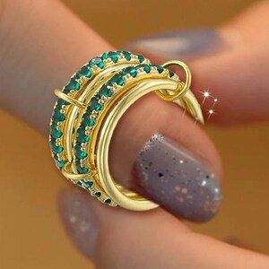 Puede incluir: Un anillo de varias bandas en tono dorado que presenta tres aros interconectados. Dos de las bandas están incrustadas con pequeñas piedras redondas de color verde vibrante, mientras que la tercera es un aro de metal dorado pulido.