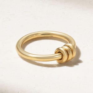 Op de afbeelding: Een goudkleurige ring met een dikke band en drie verbonden, afgeronde accentstukken. De ring is gemaakt van massief, mat metaal en ligt op een wit oppervlak. De ring is een sieraad.