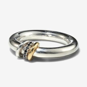 Pode incluir: Um anel prateado com uma banda espessa. O anel apresenta uma banda dourada e uma banda com pedras pretas. Uma banda prateada menor também está presente, criando um efeito em camadas. O anel está sobre um fundo branco.