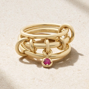 Peut inclure: Une bague dorée au design unique et lié. La bague présente plusieurs bandes imbriquées et une petite pierre précieuse rouge suspendue. La bague est présentée sur une surface neutre, soulignant ses détails complexes et sa finition polie.