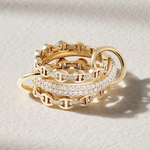 May include: A collection of gold rings with various designs. Some rings feature a chain-link pattern, while others are encrusted with small, sparkling stones. One ring has a large, circular design. The rings are displayed on a white surface.