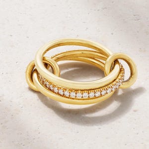Op de afbeelding: Een gouden ring met een gelaagd, in elkaar grijpend ontwerp. De middelste band is versierd met een rij kleine, heldere, rondgeslepen stenen langs de omtrek, terwijl de omliggende gladde gouden banden een gelaagd effect creëren.