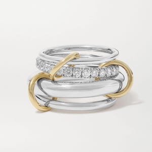May include: A stack of silver and gold rings. One ring is encrusted with small, clear gemstones. The rings are interlocked with gold connectors. The rings are displayed on a white background.