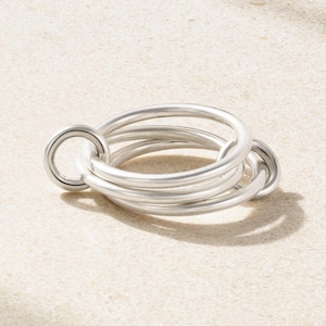 May include: A silver-colored ring composed of multiple interlocking circular bands. The ring has a smooth, metallic finish and a modern, minimalist design. The ring is shown on a neutral, light-colored surface.