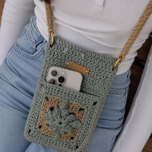 Puede incluir: Un bolso bandolera de ganchillo para teléfono en color verde salvia con un motivo cuadrado de centro beige y un diseño floral verde salvia. El bolso tiene una correa trenzada gruesa de color beige, anillas de metal dorado y una etiqueta Ciomadi.