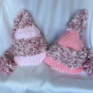 May include: Two handmade, light pink and white crochet hats with long, dangling tassels. The hats feature a textured, fluffy trim and a pointed top, perfect for a whimsical look. The hats are displayed on a white background.
