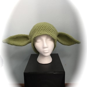 May include: A green knitted hat with large, pointed ears, resembling a character from a popular science fiction franchise. The hat is displayed on a white mannequin head, set against a light blue wall and a black surface.