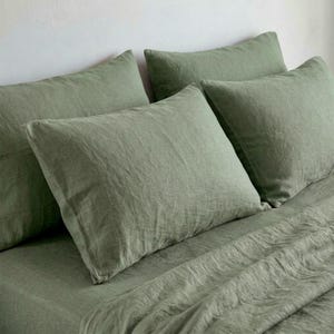 May include: Sage green linen bedding set featuring four pillowcases and a fitted sheet. The fabric has a textured finish, and the pillows are arranged on the bed. The overall aesthetic is simple and natural, perfect for a bedroom.