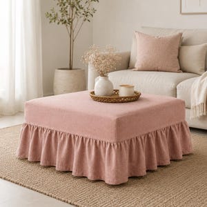 May include: A square ottoman upholstered in dusty pink fabric with a gathered ruffle skirt around the base. A round woven tray sits on top, holding a white ceramic vase filled with dried flowers and a small white cylindrical candle holder.