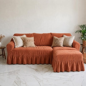 May include: An orange linen-look sectional sofa with a ruffled skirt and several decorative cushions. The sofa has a chaise lounge on the right. The cushions are in neutral tones of beige and brown. The room has a white wall and marble tile floor.