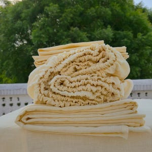 May include: A pile of cream-coloured bed linen, including a fitted sheet with elasticated edges and several folded flat sheets. The sheets are made of a soft, natural fabric, and are neatly arranged on a white surface. Green foliage is in the background.