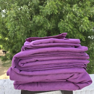 May include: A pile of folded, solid purple bed linen. The linen is neatly arranged, showing the texture and colour of the fabric. A blurred green tree forms the background.