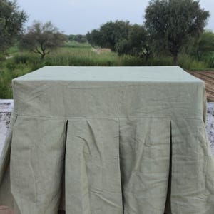 Op de afbeelding: Een saliegroene tafelkleed met een plat bovenoppervlak en een geplooide rok die vanaf de randen naar beneden hangt. De stof heeft een zichtbare textuur en een matte afwerking, wat zorgt voor een gestructureerde val rond de rechthoekige tafelvorm.