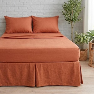 Può includere: Un letto con biancheria da letto color terracotta, tra cui un lenzuolo con angoli, un lenzuolo piano, federe e una gonna da letto. Il letto è fatto. La stanza ha una palette di colori neutri con un muro di mattoni bianchi e pavimento in legno.