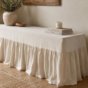 May include: A rectangular bench covered in a long, off-white fabric slipcover featuring a gathered ruffle skirt that extends to the floor. The bench is topped with a stack of two books and a small candle in a glass jar.
