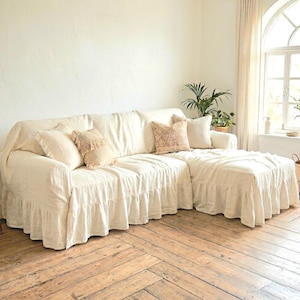 Puede incluir: Un sofá seccional color crema con una falda con volantes y varias almohadas decorativas. El sofá está en una habitación luminosa con pisos de madera y una ventana grande. Las almohadas tienen flecos y diseños estampados.