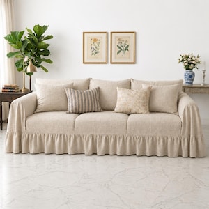 Peut inclure: Un canapé trois places beige avec une base à jupe à volants, comprenant deux coussins carrés beiges unis, un coussin rayé gris et beige, et un coussin beige à motif floral, le tout recouvert d'un tissu texturé.