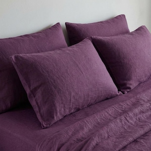 May include: A set of four plum-coloured pillowcases and a matching sheet. The bedding has a textured appearance, suggesting a linen or similar fabric. The pillows are arranged on top of the sheet, creating a layered effect.