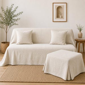 May include: A beige sofa covered with a textured fabric slipcover, featuring two matching square throw pillows. A matching square ottoman with a pleated skirt sits in front of the sofa, all placed on a woven tan area rug.