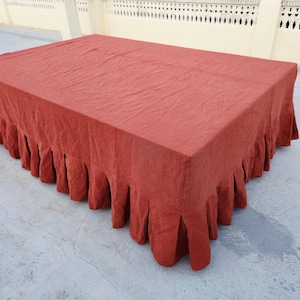 Peut inclure: Une nappe rectangulaire en lin de couleur rouille dotée d'une jupe plissée qui retombe sur les bords. Le tissu présente un tissage texturé visible et recouvre le dessus ainsi que les côtés d'une surface, créant un aspect drapé et superposé.