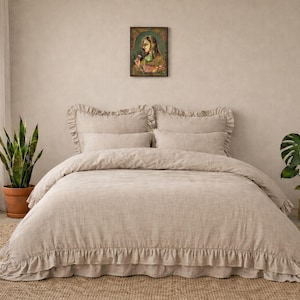 May include: A neutral-toned linen bedding set with frilled edges, including a duvet cover and pillows, is presented on a bed. The room has a muted colour scheme, with plants in terracotta and gold pots, and a framed artwork above the bed.