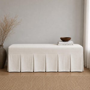 May include: A rectangular off-white upholstered bench featuring a pleated skirt base. On top of the bench sits a dark brown wooden bowl and two stacked books with the visible text "KINFOLK" and "LIVE BEAUTIFUL" printed on their spines.