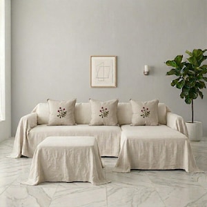 May include: A beige sectional sofa and matching ottoman covered in a light-colored fabric. Three square throw pillows are arranged on the sofa, each featuring a small, centered floral embroidery of red flowers with green stems and leaves.