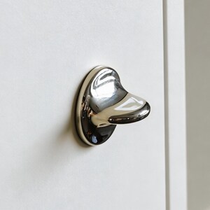 Può includere: Una maniglia per armadio cromata lucida a forma di becco d'anatra è montata su una porta di un armadio bianco. La maniglia ha una piastra posteriore arrotondata e una superficie liscia e riflettente, che aggiunge un tocco di eleganza all'ebanisteria.