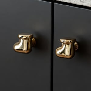 May include: Two gold-coloured cabinet knobs shaped like a baby bootie are mounted on a dark grey cabinet door. The knobs have a shiny, reflective surface. A light-coloured countertop is visible in the background.