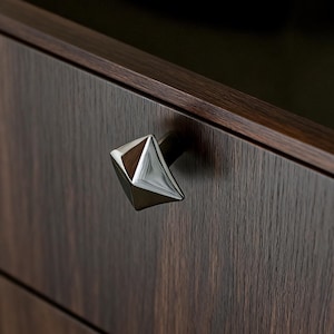 May include: A close-up of a dark brown wooden cabinet with a geometric, silver-coloured drawer pull. The drawer pull is a faceted, diamond-shaped design. The wood grain is visible on the cabinet's surface, adding texture to the piece.