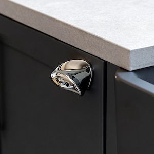 May include: A chrome cabinet pull with an unusual, flowing form. It's fixed to a dark grey cabinet door, with a light grey countertop above. The design is contemporary and minimalist, suitable for modern kitchens.
