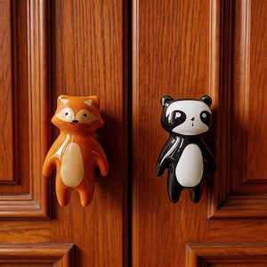 Può includere: Due pomelli per armadi a forma di animali. Uno è una volpe arancione con pancia color crema, l'altro un panda bianco e nero. Entrambi sono attaccati a una porta di un armadio in legno.