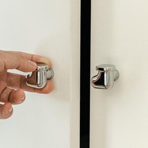 Pu&ograve; includere: Pomelli cromati lucidi per armadi con un design unico e arrotondato. Una manopola viene azionata da una mano, mentre un'altra &egrave; fissata a un'anta bianca. I pomelli hanno un'estetica moderna e minimalista.