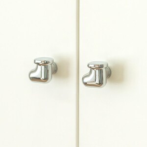 Pu&ograve; includere: Due pomelli cromati lucidi a forma di calzino sono montati su un'anta di un armadio color crema. I pomelli hanno una superficie liscia e riflettente, che aggiunge un tocco moderno al mobile.